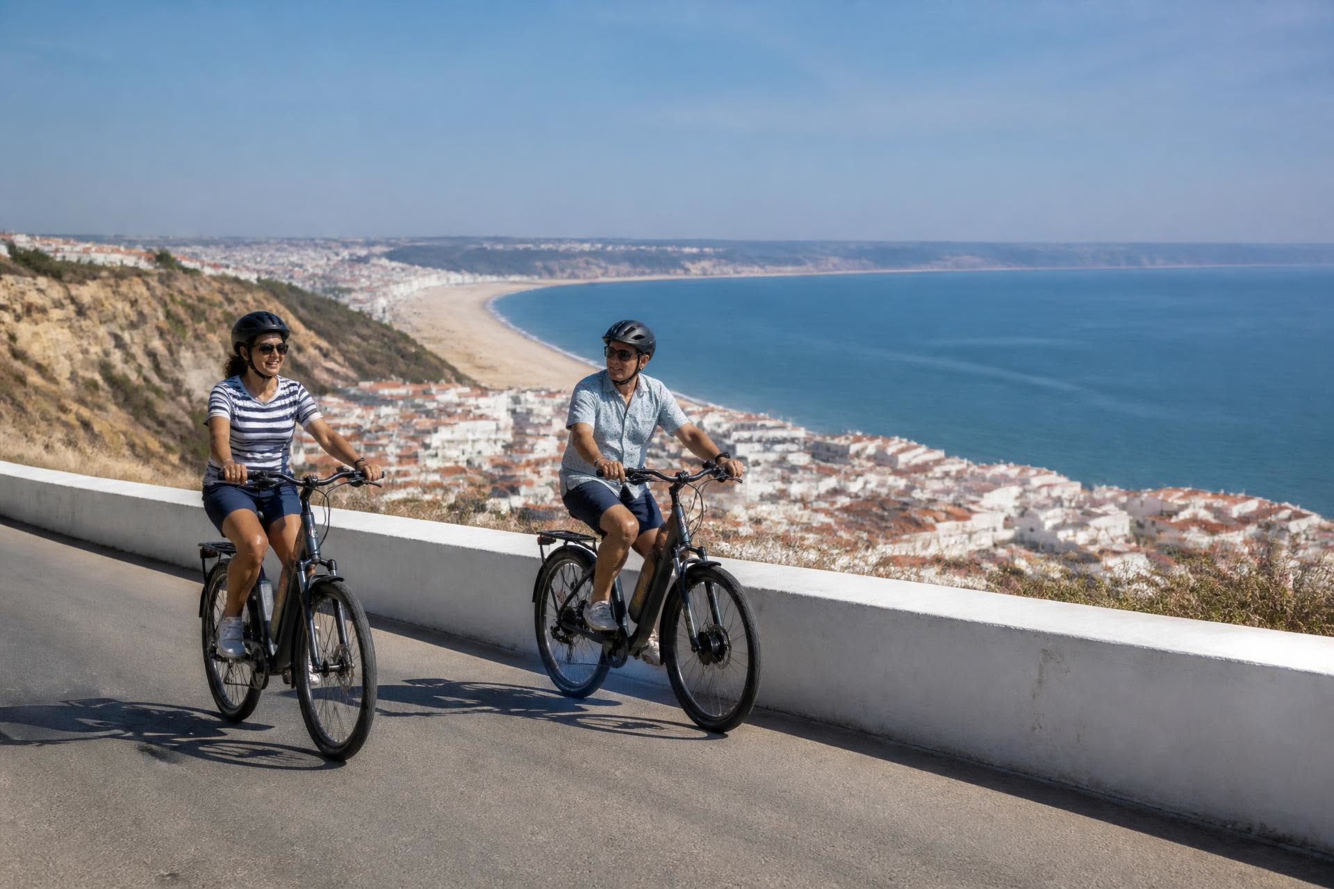 Two cyclists riding electric bikes on the clifftop road above Nazaré town beach